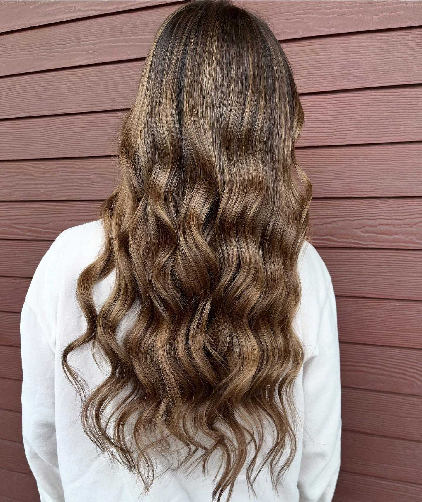 Wavy brown hair with blonde highlights cascading down a person's back against a wooden wall.