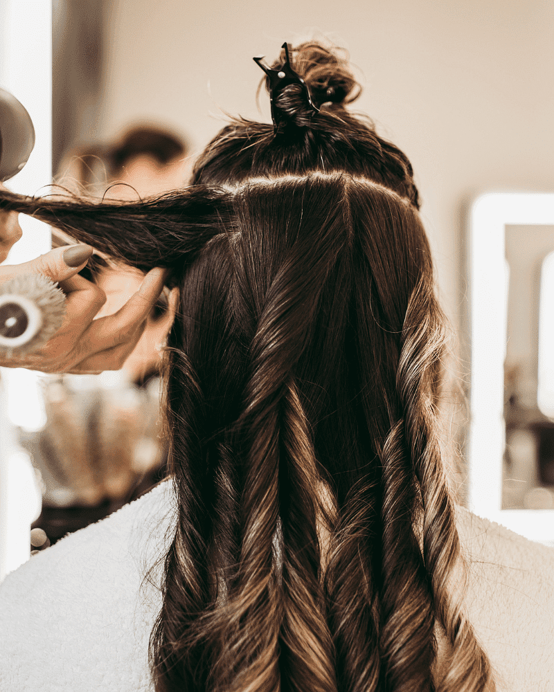 Stylist curling a client's long hair in a salon, using a round brush and a blow dryer.