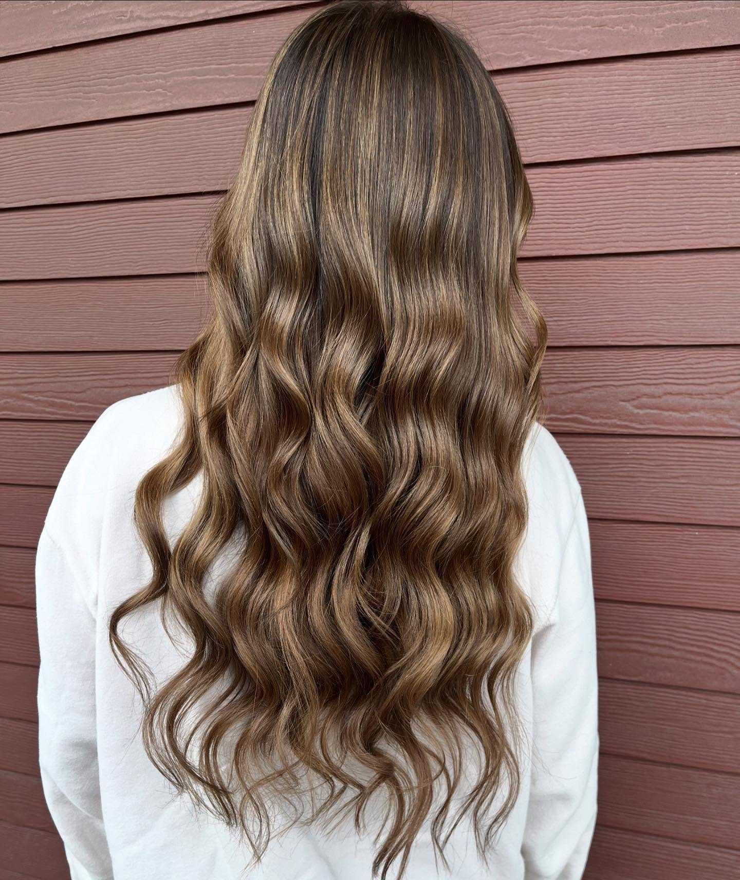 Wavy brown hair with blonde highlights cascading down a person's back against a wooden wall.