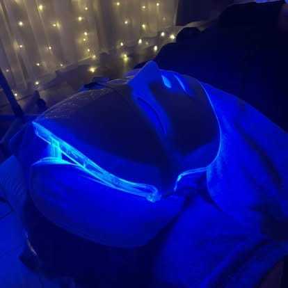 Person undergoing LED light therapy with a blue illuminated mask in a dimly lit room.